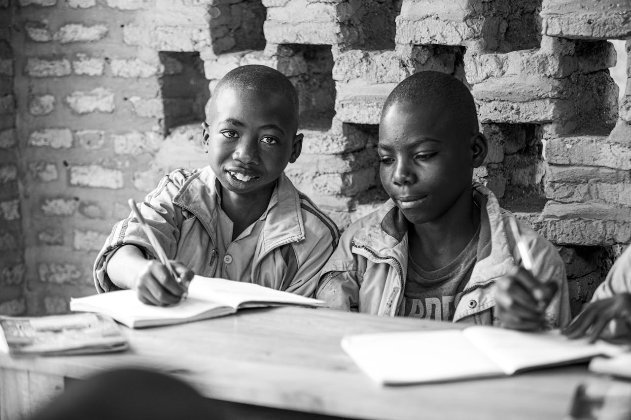 hero-img-02 Black and white photo of two children studying with notebooks and pens against a brick wall.